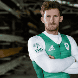 Irish Rower and Olympic Silver Medallist Gary O'Donovan standing in front of rowing boats in his Ireland Olympic uniform